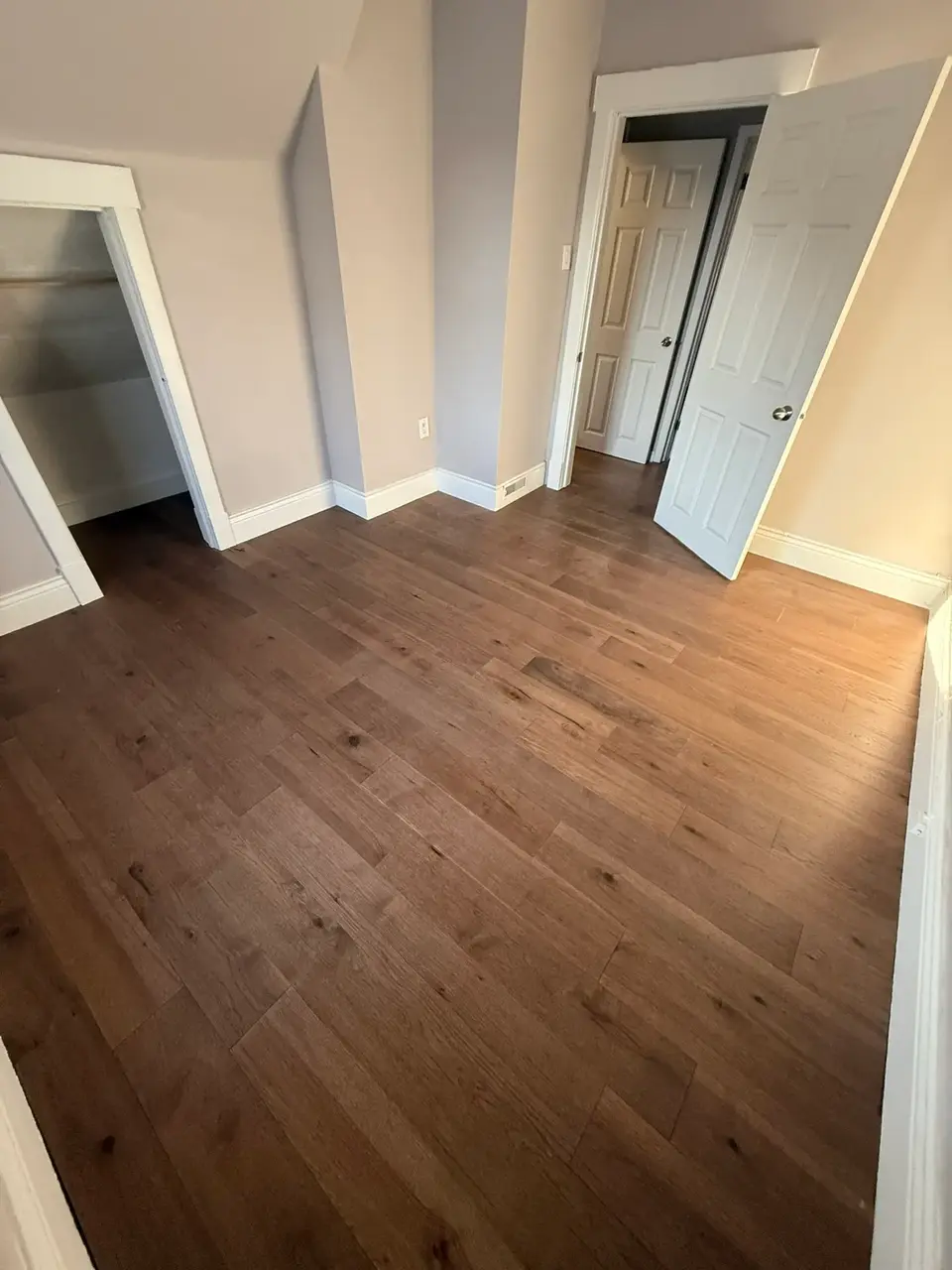 Warm oak hardwood flooring installed in a compact bedroom with angled walls, white baseboards, and natural light coming through the doorway.