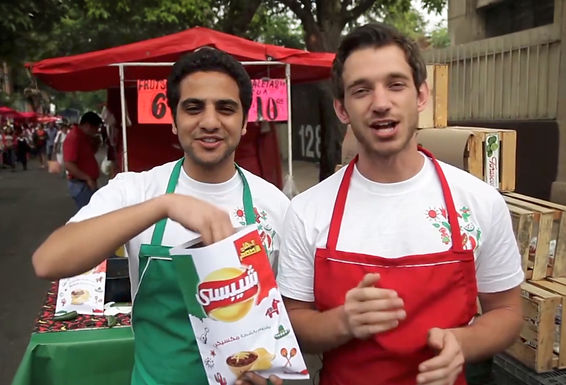 Two young men wearing white t-shirts with Mexican-themed embroidery and colorful aprons—one green and one red—stand in front of a street market stall with red tents. They are smiling and engaging with the camera while holding a bag of Lay’s chips with Arabic text and Mexican-themed branding. The background is filled with people shopping, food stands, and crates, creating a lively market atmosphere.
