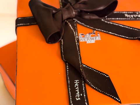 Sculpted cake resembling a Hermès orange gift box with an edible black bow and ribbon, showcasing intricate craftsmanship