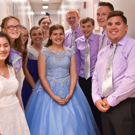 A group of young people, likely teenagers, dressed in formal attire, poses in a hallway. The young women are wearing various light-colored dresses, and the young men are wearing light purple shirts and ties with dark pants.