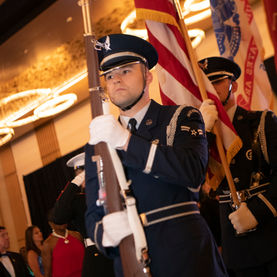 Two individuals in military honor guard uniforms are carrying rifles and flags, including the American flag, during an indoor event. People in formal wear are visible in the blurred background.