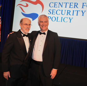 Two men in black tuxedos and bow ties are smiling and standing with their arms around each other on a stage. 