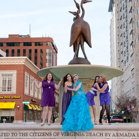 A group of five young women in formal dresses poses around a large fountain with a bird sculpture in a city setting. One woman in the center is wearing a bright blue ruffled ballgown and a tiara, while the others are wearing shorter purple dresses. Buildings are visible in the background.