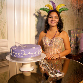 A young woman wearing a formal corset-style top, a purple beaded necklace, and a festive headpiece poses with a purple cake decorated with white swirls and a butterfly. 
