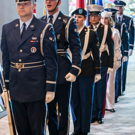 A line of individuals in military honor guard uniforms stands indoors, holding flags including the US flag and what appear to be service branch flags.