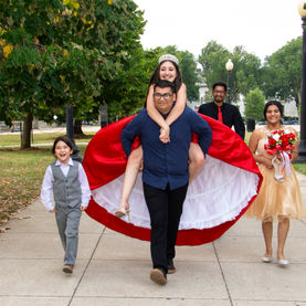 A group of four people walks along a paved path outdoors. A young woman in a red quinceañera dress and tiara is getting a piggyback ride from a man in glasses and a blue shirt. 