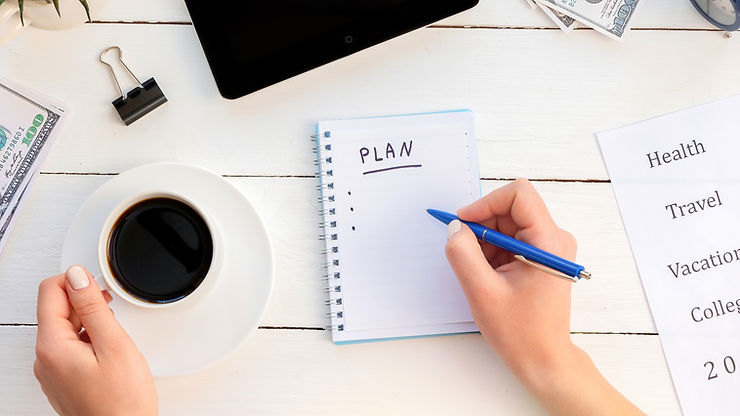 Person writes "PLAN" in notebook, holding a pen and coffee. Nearby are cash, a tablet, and a list with "Health, Travel" notes on a white table.
