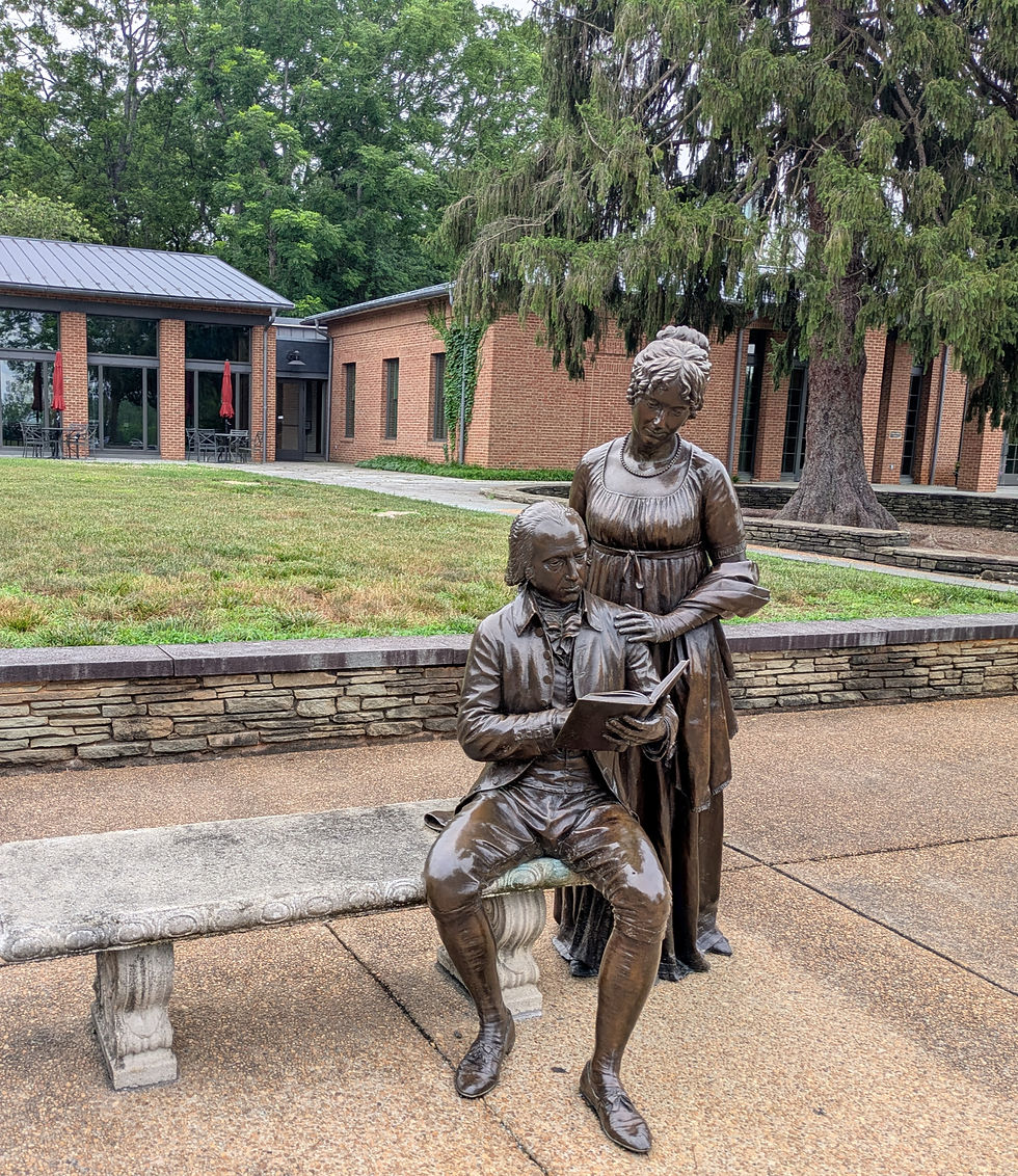 Bronze statue of a seated James Madison reading while a Dolley Madison stands behind him, in a courtyard with brick buildings and tall trees.