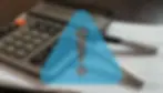 Calculator and pen on spreadsheets with a blue warning triangle overlay. Dark, professional setting.