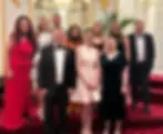 Amy and team in formal attire pose on a red carpeted staircase. Elegant setting with ornate gold details and chandeliers. Smiling faces.