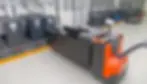 Orange electric pallet jack charging large black batteries in an industrial setting. Gray floor and wall; organised cables above.