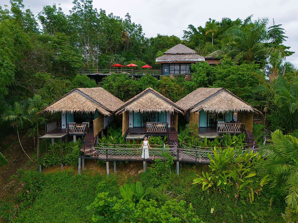 Woman stands on a deck below three thatched bungalows in a lush tropical hillside resort, surrounded by dense green jungle.