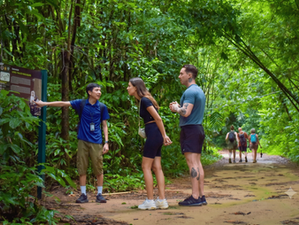 Guide points to a Long-tailed macaques sign as two tourists listen on a lush jungle trail, with hikers in the background.