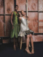 Two women in stylish dresses; one in green standing, the other in metallic silver seated on a stool. Wooden backdrop, elegant mood.
