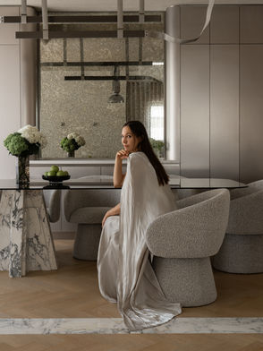 Interior designer seated at a marble and glass dining table with sculptural grey boucle chairs, wearing a flowing silver gown, in a contemporary neutral-toned dining space with herringbone wood floors and fresh hydrangea arrangements.