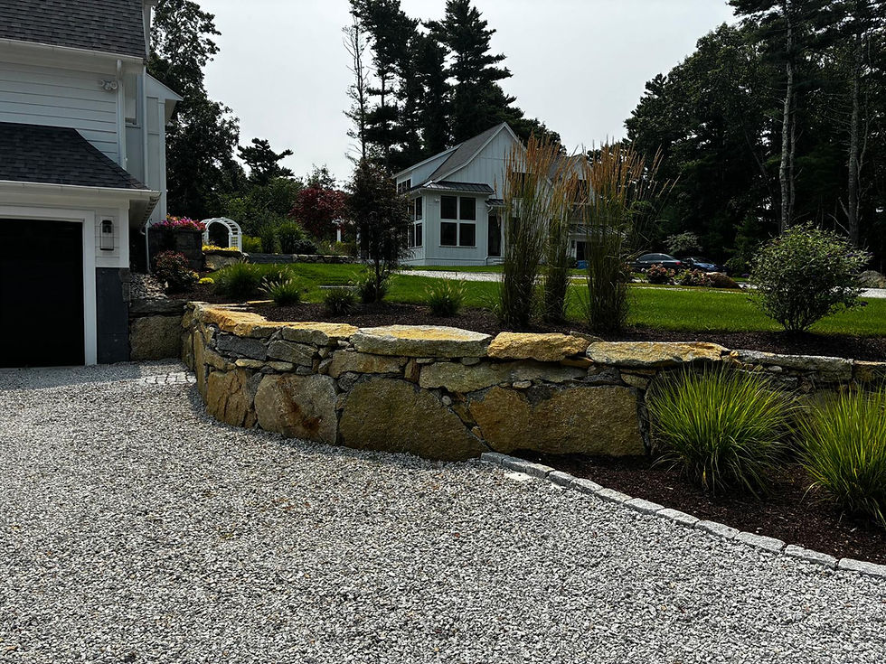A house with a stone wall and gravel driveway, surrounded by grass and trees. White pergola visible, creating a serene garden setting.