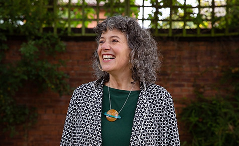 A laughing woman in her 40s stands in front of a garden wall. She has curly brown/grey hair and a necklace with a planet on it.