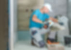 Contractor in blue shirt and white cap applying adhesive and installing gray ceramic tiles in shower area.