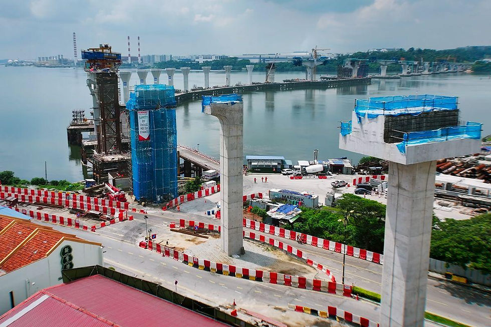 Johor-Singapore RTS construction over water with tall pillars. Red and white barriers line the road. Blue scaffolding on structures. Overcast sky.