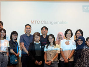 Group of 12 people smiling indoors in front of a screen displaying "MTC Changemaker." Casual attire, diverse expressions, warm lighting.
