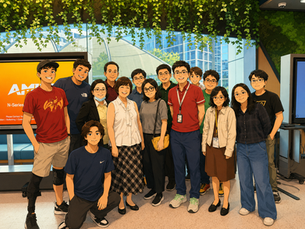 A group of smiling people pose indoors with plants hanging above. There’s a screen displaying “AMD” in the background. Casual attire.