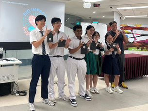 Group of seven people, mostly in school uniforms, smiling and holding notebooks in a room with a screen displaying "Big Impact-Bigger Hearts."