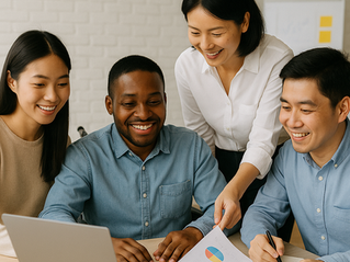 Four people smile while discussing charts at a table with a laptop. Bright, casual setting with a white brick wall background.