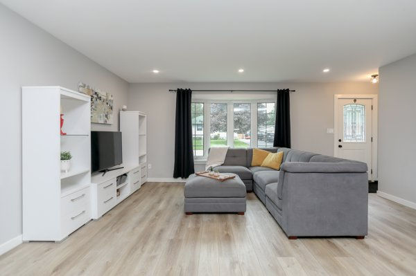 Bright modern living room with gray sectional, ottoman, white TV shelves, black curtains, and front door by large window representing a finished living room and open-concept main floor after renovation