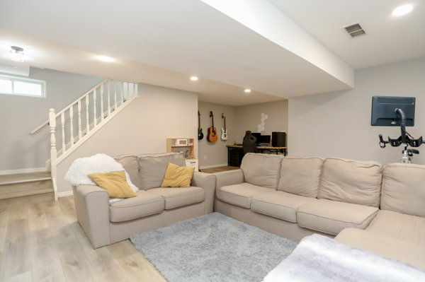 Bright basement family room after home renovation with beige sectional, gray rug, stairs, desk with guitars, and a computer nook.