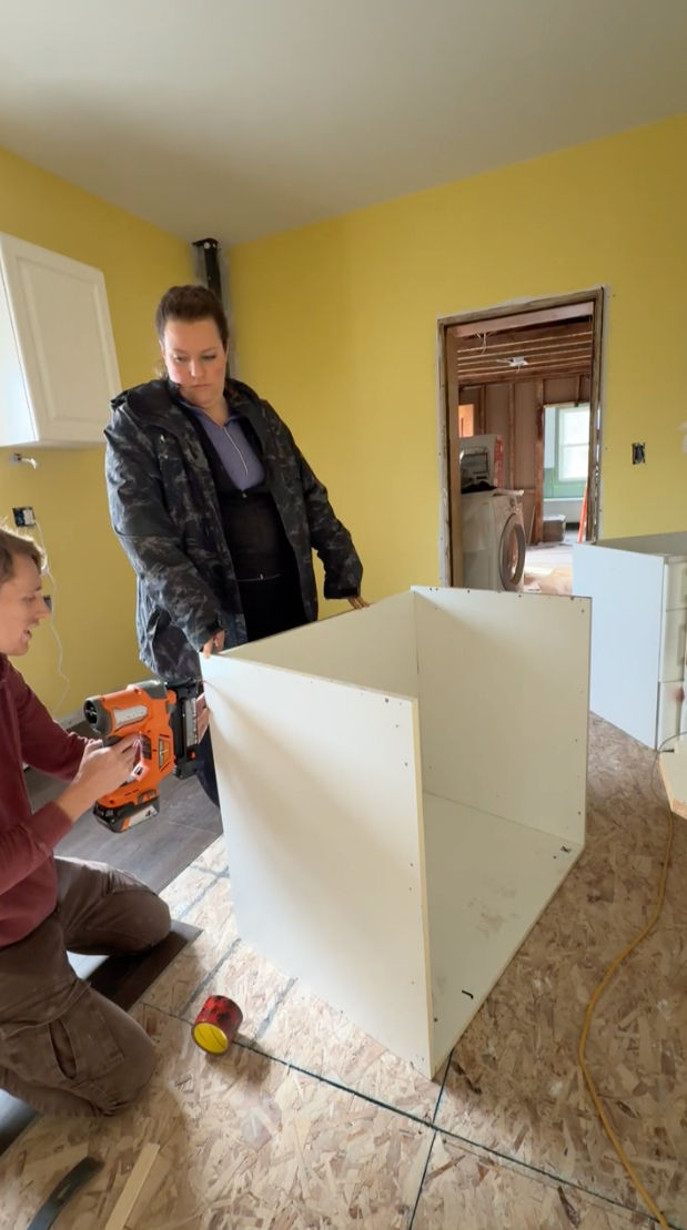 A Canadian DIY reno couple assemble a white cabinet box in a yellow-walled kitchen, one using an orange nail gun.