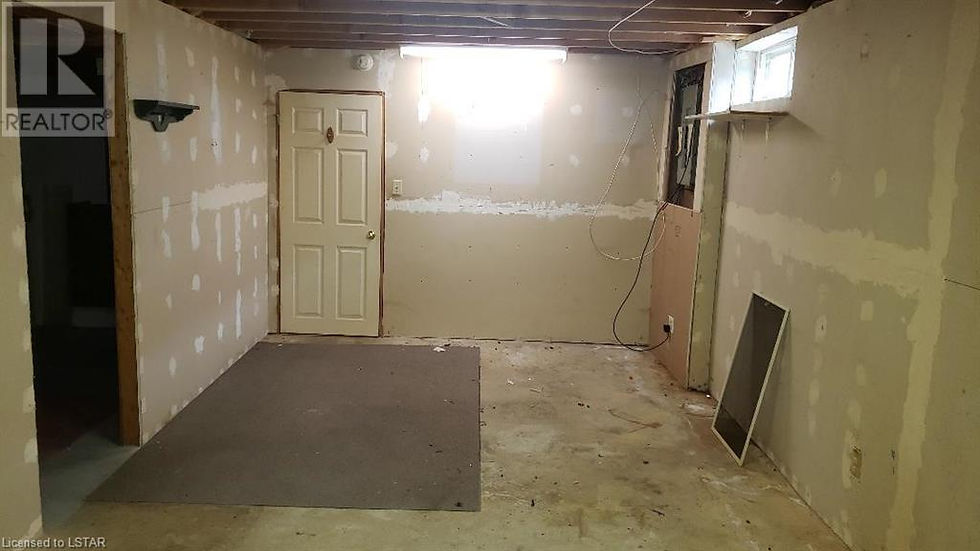 Unfinished basement space before bedroom renovation with patched drywall, white door, exposed beams, small window, dark opening to unfinished laundry room on left and closed bedroom door in centre of the photo.