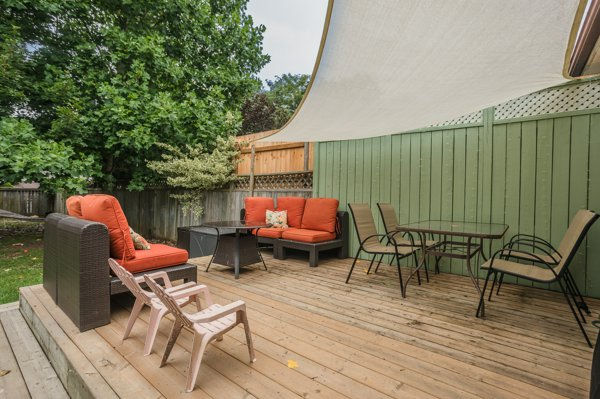 Finished DIY wooden deck after our home renovation with orange-cushioned patio seating, table and chairs under a shade sail, beside a green fence and trees.