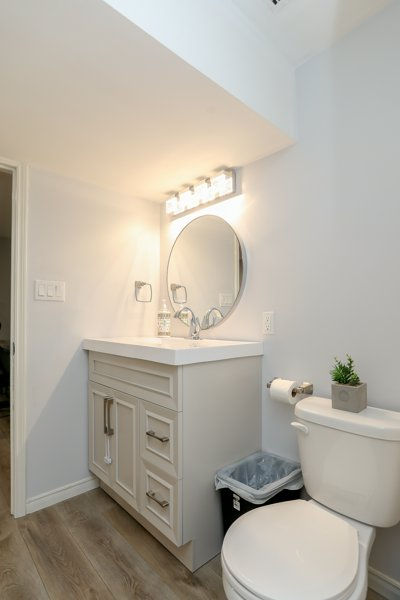 Updated basement bathroom after simple DIY renovation turning the room into a bright modern bathroom with white vanity, round mirror, toilet, and small plant beside a trash bin.