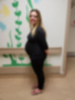 A smiling pregnant woman in black clothing stands against a wall with green seaweed and colourful fish designs in the labour and delivery area waiting for induction.