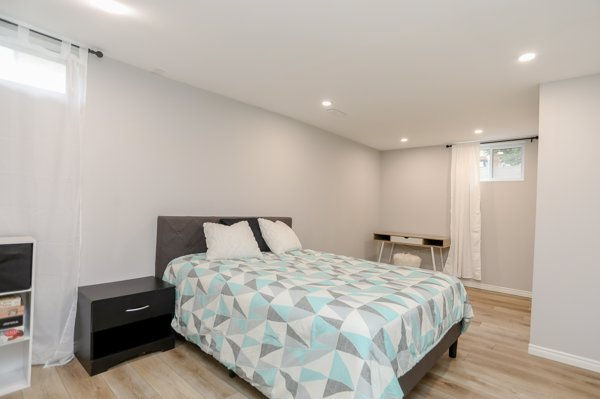 Legal basement bedroom after finishing the previously unfinished space. It' s now a bright minimalist bedroom with a geometric quilted bed, black nightstand, desk, and small window on light wood floors.