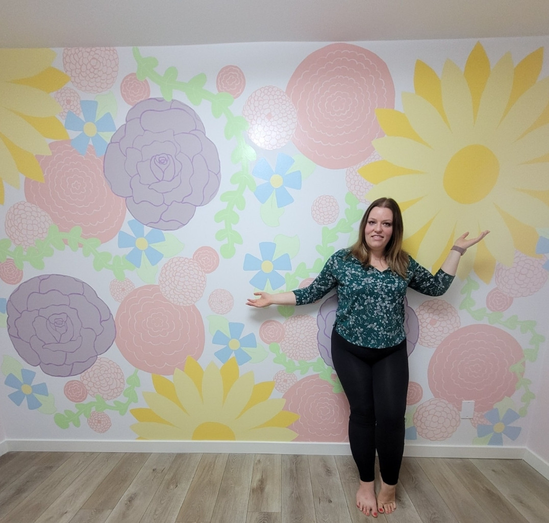 Woman posing with arms out in a bright room with hand-painted floral wall mural in daughter's bedroom.