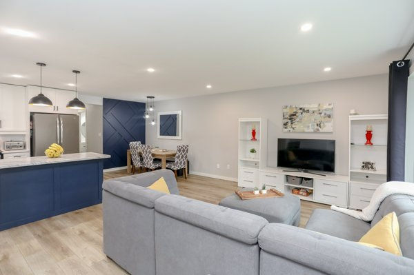Living room home renovation before and after wall removal reveals a modern open-plan living room with gray sectional, blue kitchen island, dining table, TV wall, and warm pendant lights in a bright home