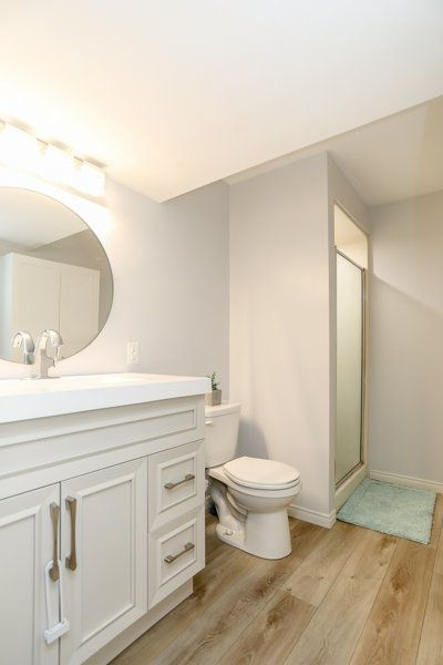 Downstairs bathroom after new flooring, vanity, paint and lighting shows a bright modern bathroom with white vanity, round mirror, toilet, glass shower, and light blue rug on wood-look floor.