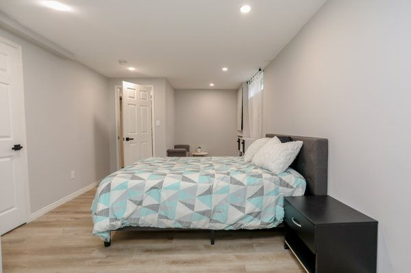 Minimal bedroom with bed in geometric quilt, black nightstand, open door, and recessed lights in a calm gray room represents a Finished basement bedroom with new closet and egress window.