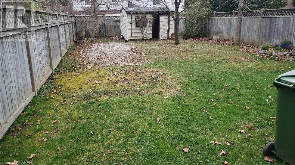 Fenced backyard with patchy grass, gravel area, shed and tree; green trash bin at right, realtor watermark visible. Backyard before relocating shed that divided the usable yard space.