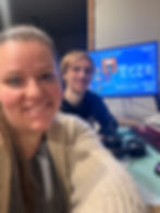 Two people smiling in front of a computer screen displaying a blueprint and inventory from "Blue Prince" video game for couple. The setting is indoors, with headphones nearby on date night.