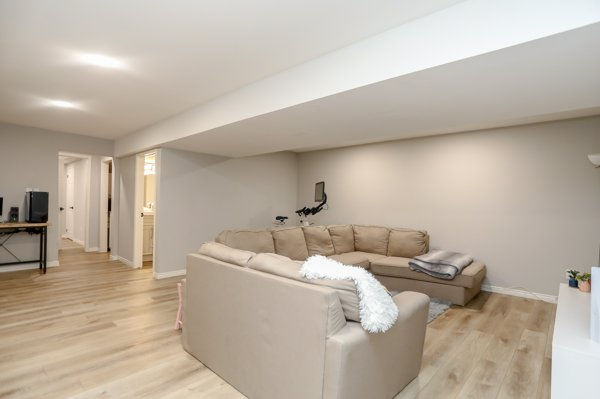 Spacious beige basement living room with sectional sofa, chair and folded blanket on wood floors, calm and tidy, no people. The basement family room after removing wood-burning furnace
