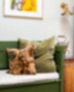 Green bench with floral and plain cushions. Wall art with yellow abstract, brass sconce above. Kettle on wooden side table. Cozy setting.