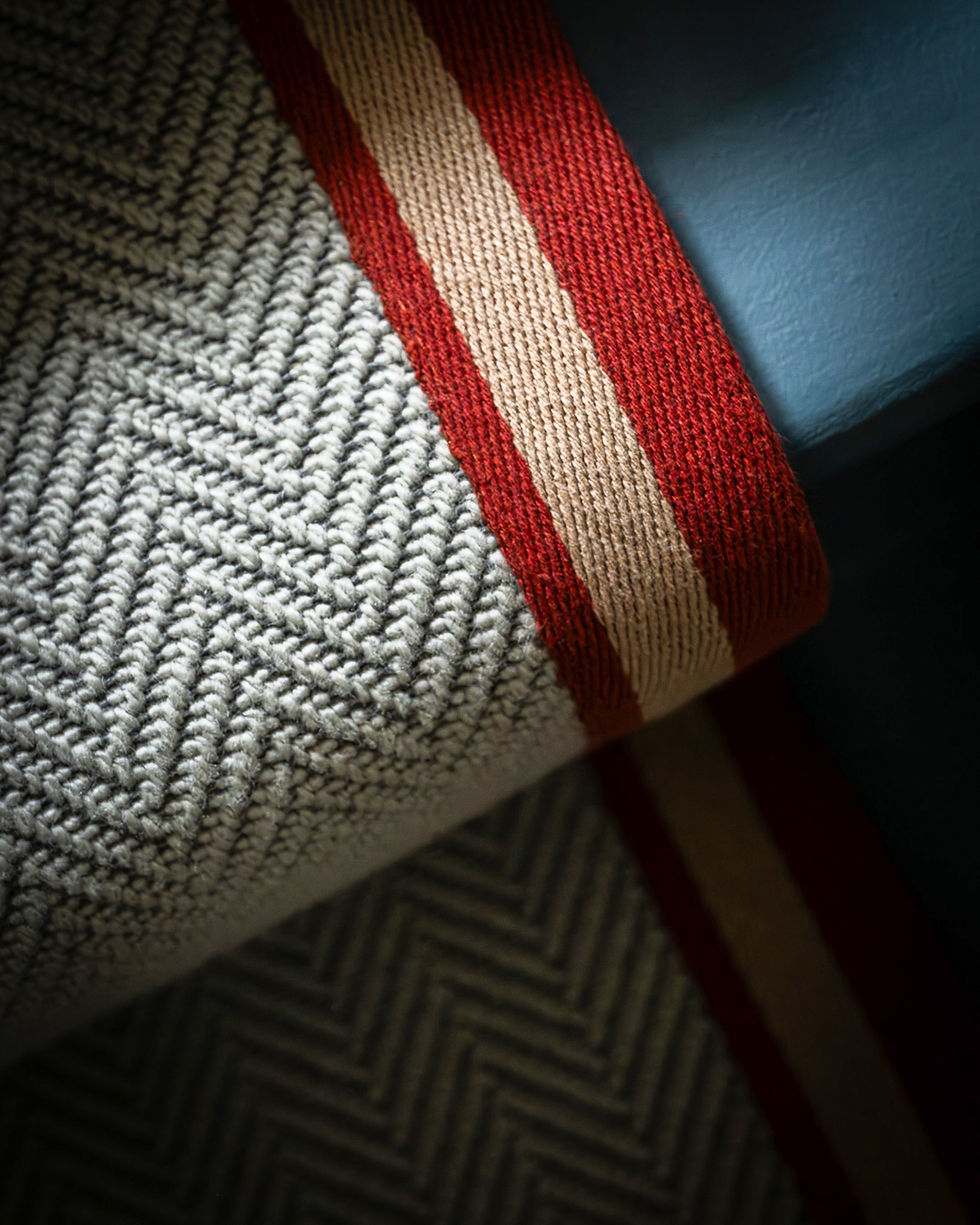Close-up of a woven gray herringbone rug with a red and beige striped edge, partially rolled on a blue surface, creating a cozy mood.