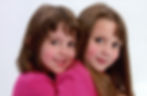 Two young girls in pink shirts smiling, embracing against a plain white background. 