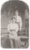 Vintage photo of a woman seated and a man standing behind her on a wooden bench. Both wear early 20th-century attire, with a neutral backdrop.