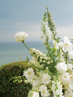 Elegant white floral arrangement with roses and delphiniums set against a scenic coastal backdrop with ocean and sky in view.