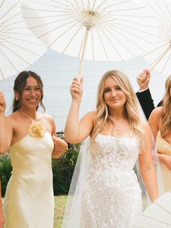 Radiant bride and her bridesmaid pose with white parasols, all smiles on a sunny wedding day.