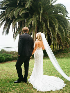 Bride and groom standing hand in hand on a grassy lawn, with a large palm tree and ocean view in the background.