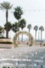 White floral arch and rows of chairs set for a wedding on a sunny beachside with palm trees and string lights overhead.